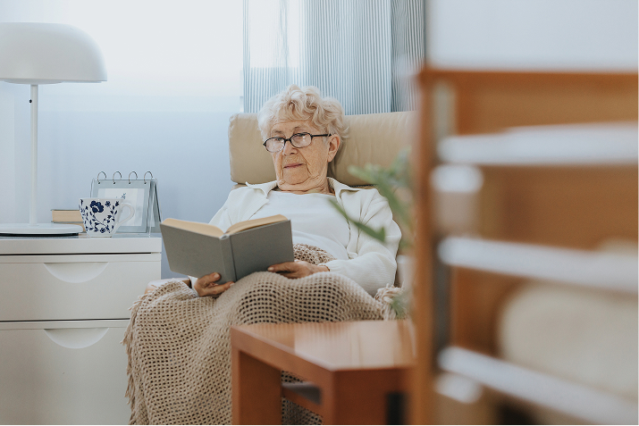 Elderly woman with glasses reads a book while relaxing in a cozy armchair under a blanket. TITLE