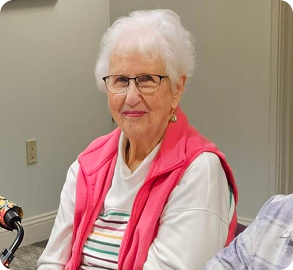 A smiling senior woman with white hair and glasses wears a pink vest over a striped