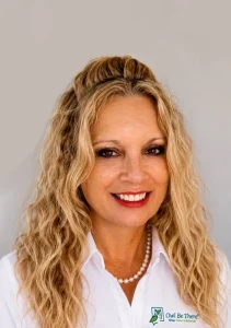 A smiling woman with long blonde wavy hair wears a white collared shirt and pearl necklace.