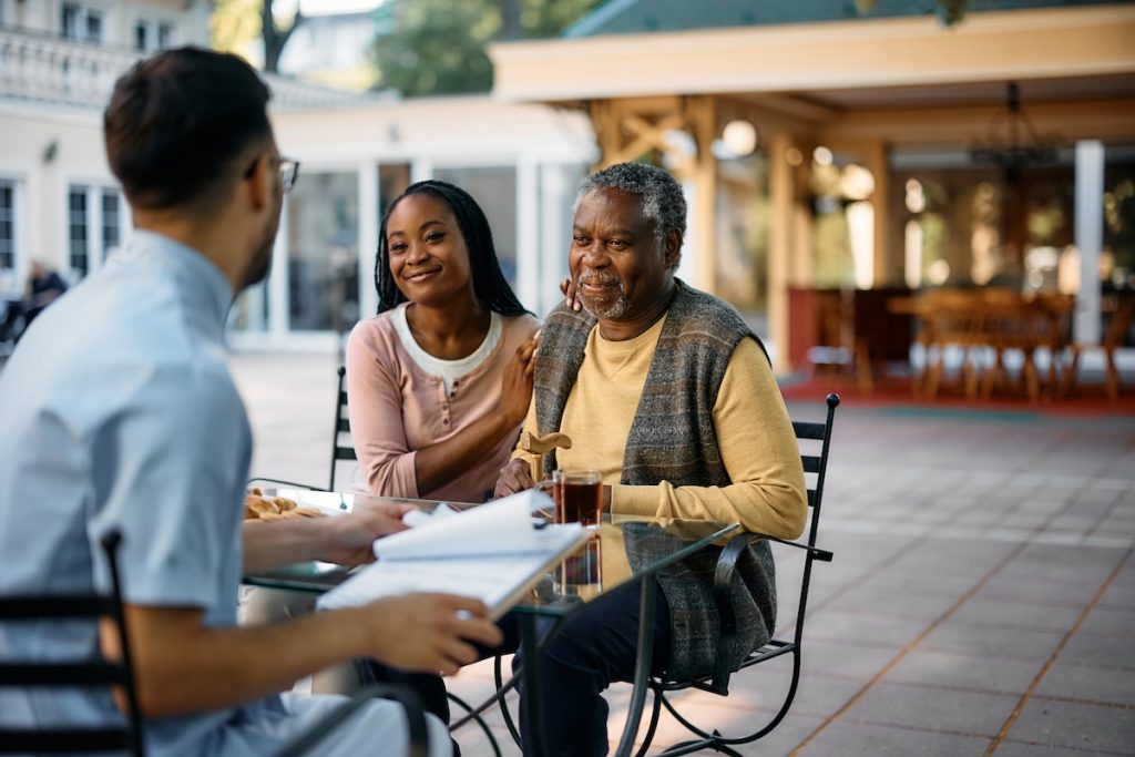 A professional discusses documents with a smiling senior man and his caregiver at an outdoor table.
