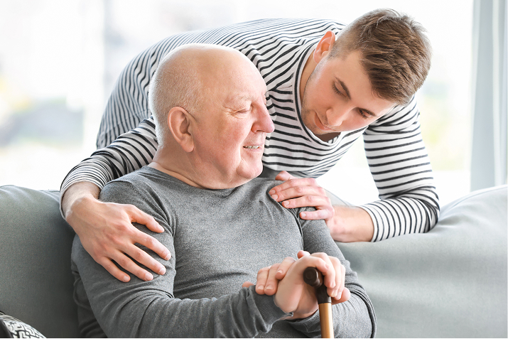 A younger man leans over an older man holding a walking stick, offering comfort and support.