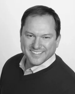 A smiling man in a dark V-neck sweater and collared shirt in a black and white headshot.