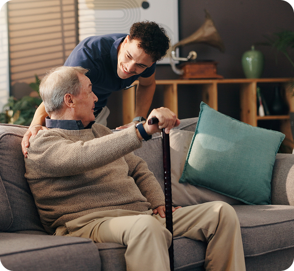 A smiling young man leans over and supports an older man holding a cane on a couch.