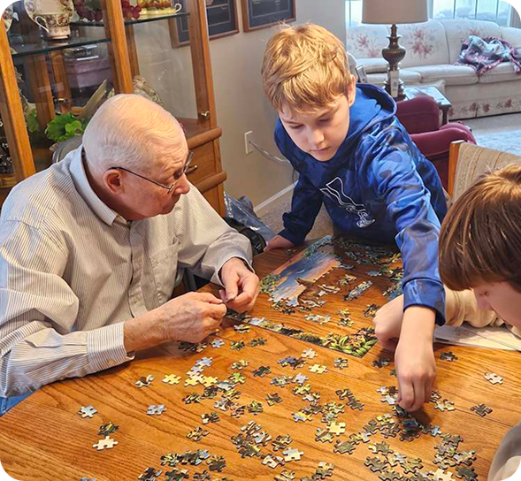 Grandfather and two boys assemble a jigsaw puzzle together at a wooden table. TITLE: