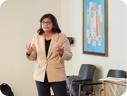 A professional woman in glasses and a tan blazer gestures while presenting.