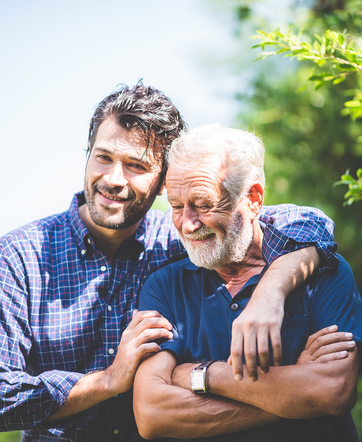 A smiling young man embraces an older man with a white beard outdoors on a sunny