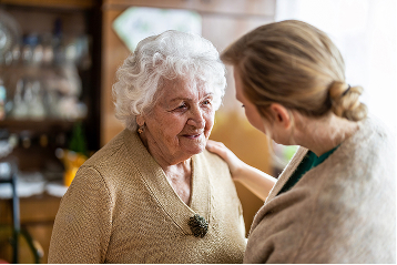 A smiling elderly woman looks at a younger woman gently touching her shoulder.