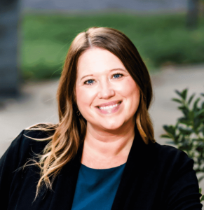 A smiling woman with long brown hair and blue eyes wears a dark blazer, leaning on a railing
