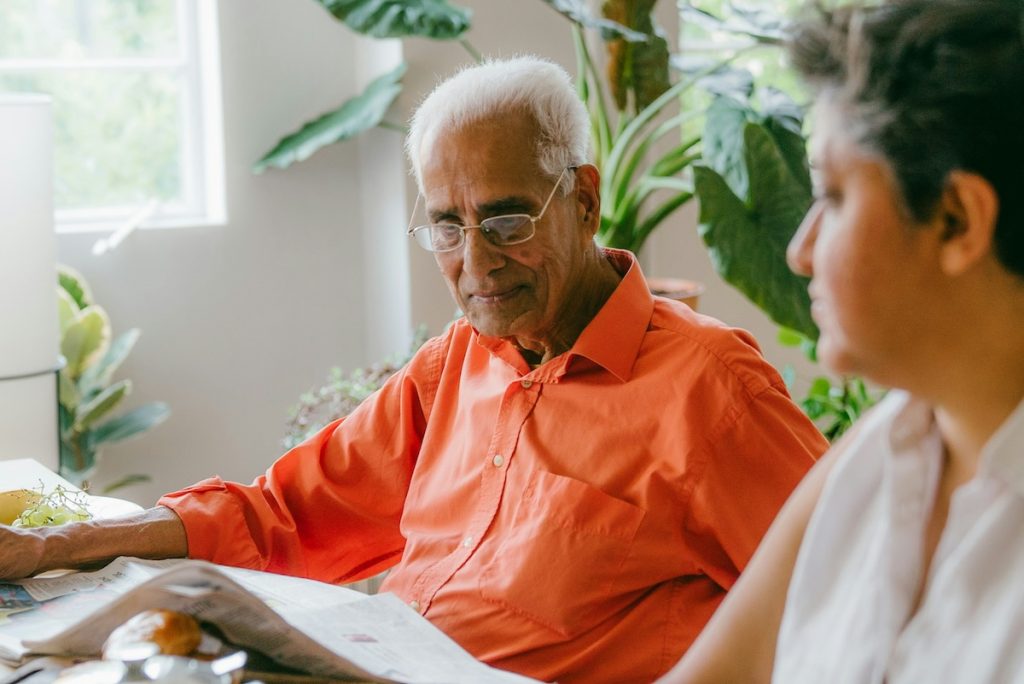 An older man with white hair and glasses reads a newspaper next to a younger person.