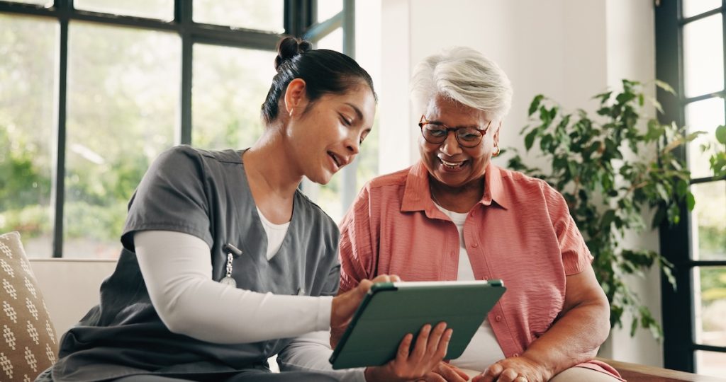 A caregiver shows a smiling senior woman information on a tablet in a bright room. TITLE