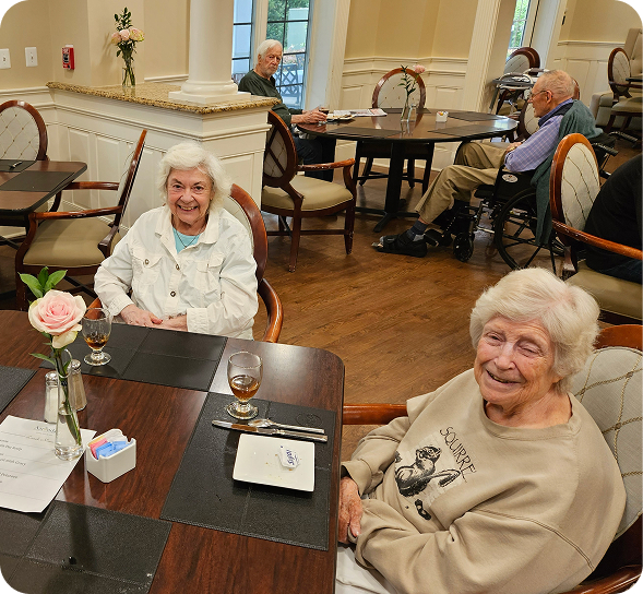 Two smiling elderly women enjoy a meal in a bright senior living community dining room.