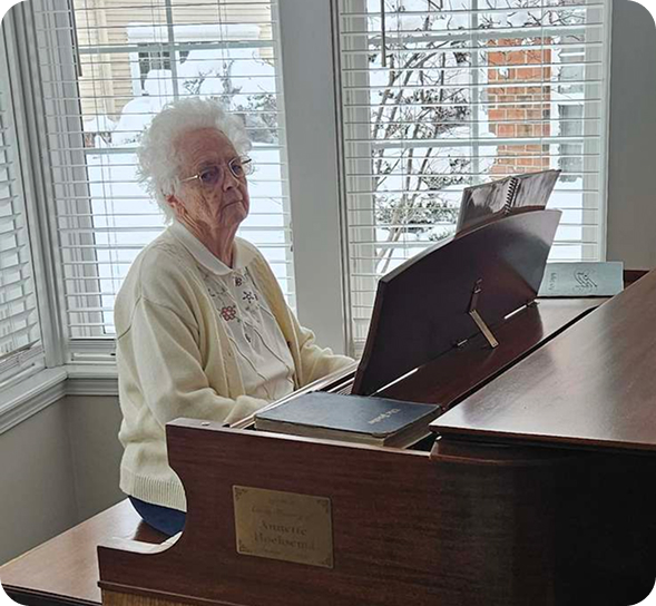 White-haired woman in glasses sits at a piano with sheet music, looking out a snowy window