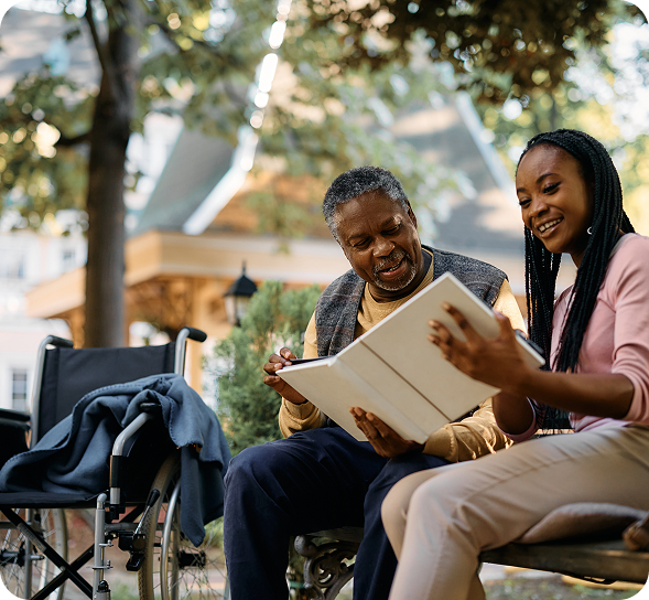 A smiling senior man and young woman share a photo album on a park bench next to a wheelchair