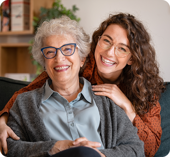 A happy older woman and younger woman with curly hair smile broadly, embracing on a couch.