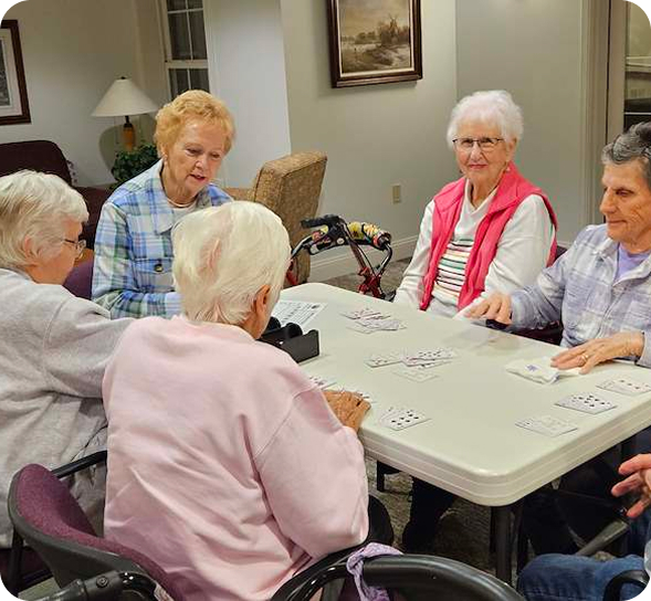 Five senior women gather around a table, playing cards and socializing in a bright room.