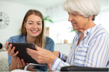 A smiling young woman helps an older woman in a wheelchair use a digital tablet.