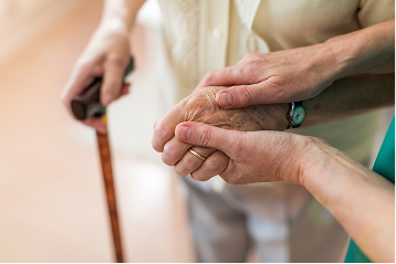 A caregiver's hand gently supports an elderly person's hand holding a walking cane.