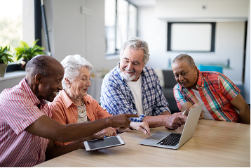 Four smiling seniors learn together using a laptop and tablet at a table. TITLE:
