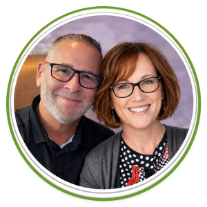 A smiling man with a beard and woman with red hair, both wearing glasses.