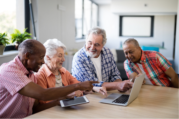 A diverse group of smiling seniors learn to use a laptop and tablet together.