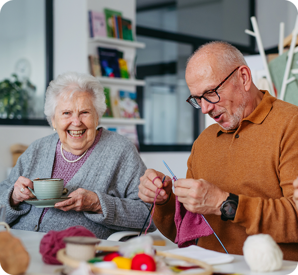 Two smiling seniors enjoy crafting together, one knitting and the other drinking tea.