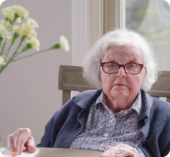 An elderly woman with white hair and red glasses sits at a table, looking seriously forward