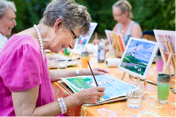 A smiling senior woman paints a lighthouse scene during an outdoor art class.