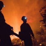 Two men are standing in front of a fire.