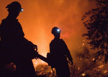 Two men are standing in front of a fire.